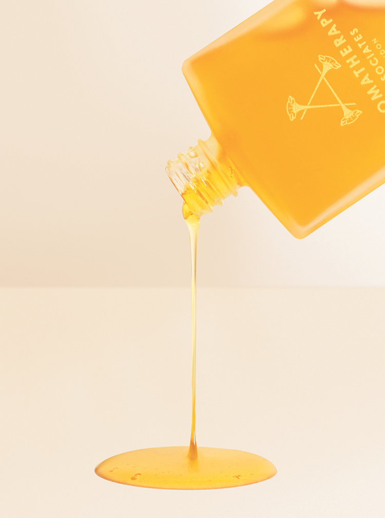 An amber-colored bottle of Omatherapy hair oil is tilted, pouring a thick, golden yellow liquid in a single stream onto a clean, light beige surface.