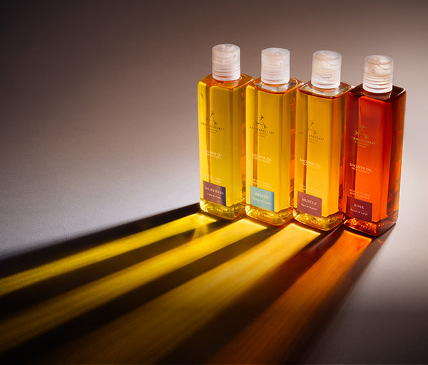 Four rectangular bottles of aromatherapy associates oil arranged in a row on a dark surface, casting long yellow and orange shadows. The bottles contain golden, amber, and reddish-orange liquids, with white caps and gold branding on the labels.